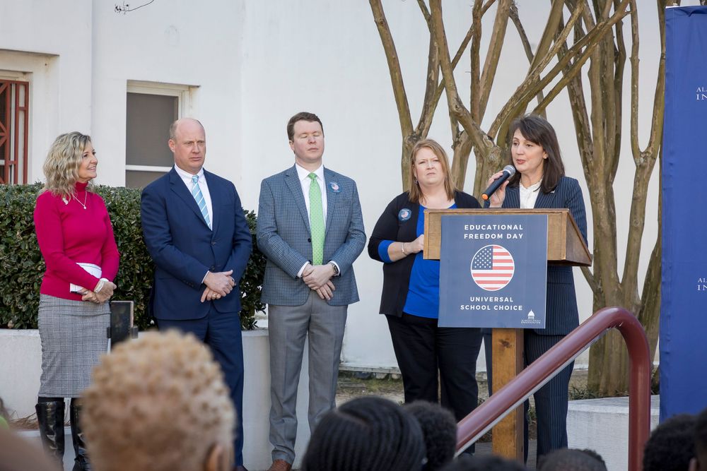Group presents at Educational Freedom Day event with white building and trees in background.