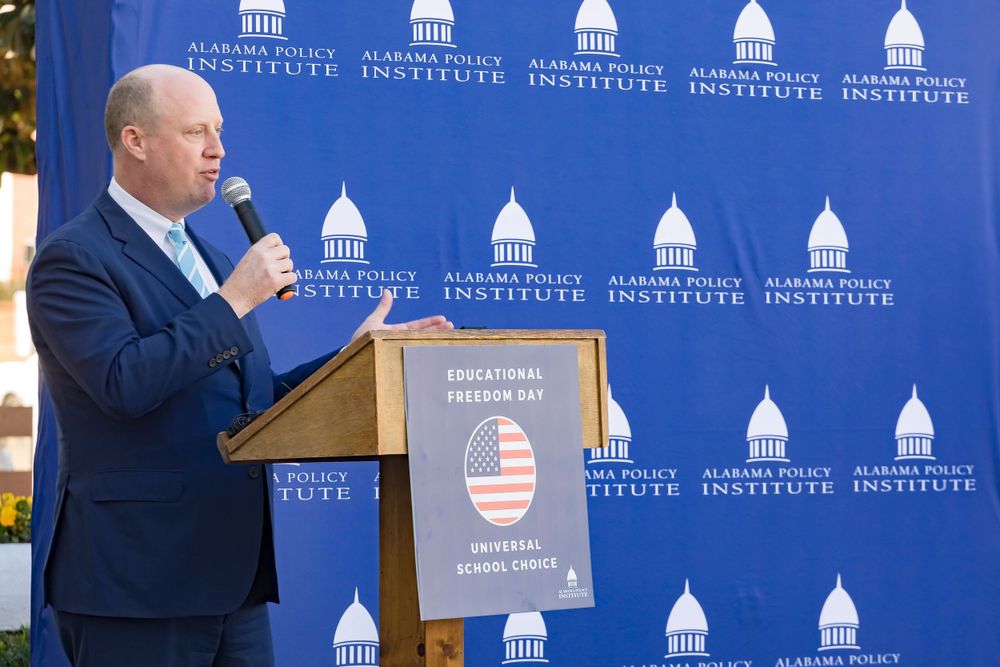 Speaker addresses crowd at podium with Alabama Policy Institute backdrop during outdoor event.