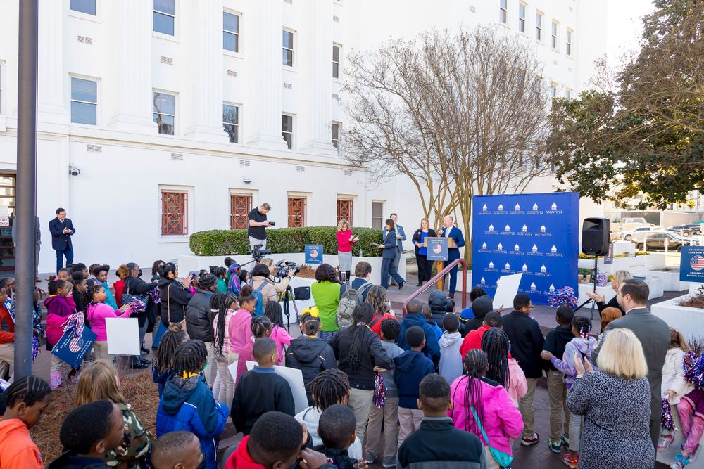 Audience listens to speaker at outdoor education event with white building and blue backdrop visible.