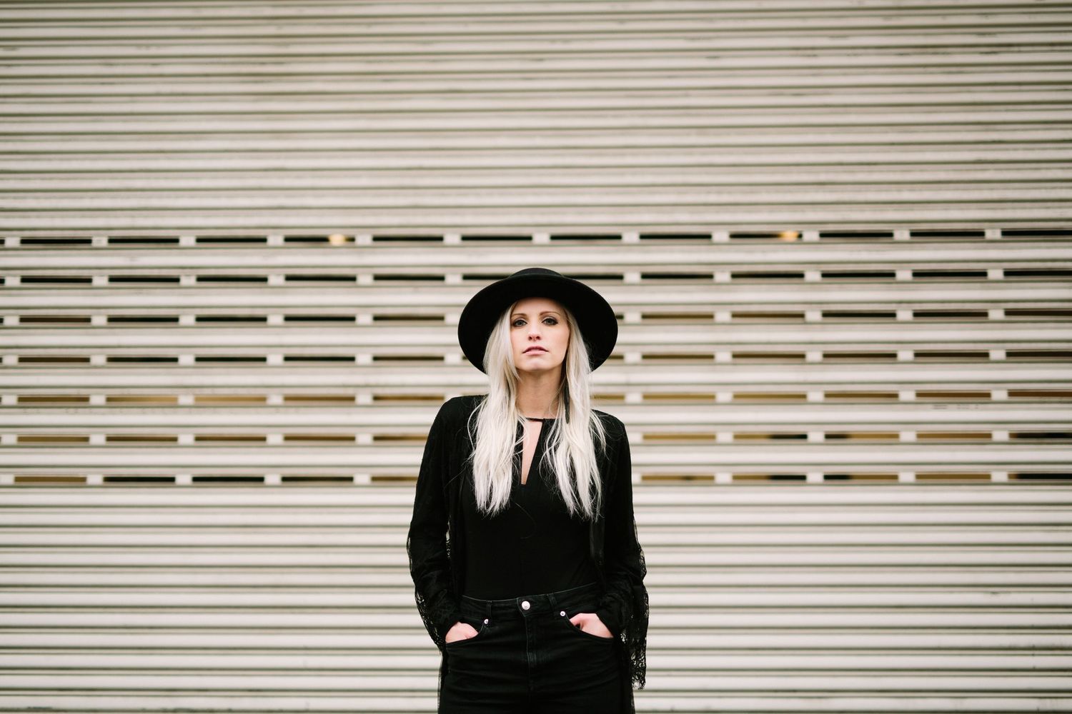 A stylish blonde woman in black outfit and wide-brim hat stands confidently in front of a metal shutter wall.