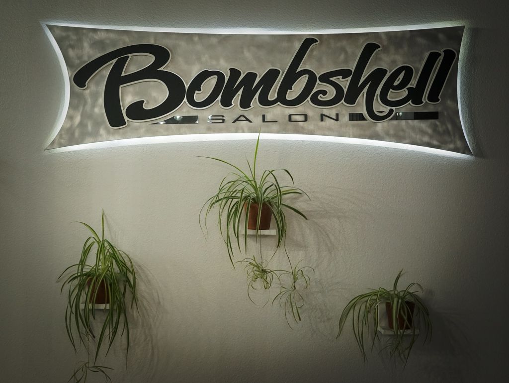 Illuminated Bombshell Salon neon sign on wall with hanging spider plants for decor.