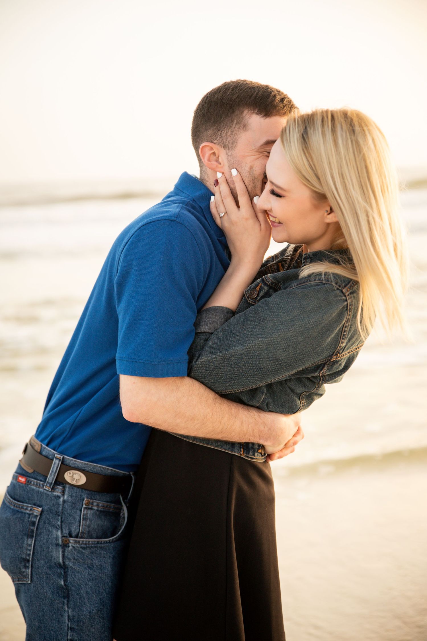Couple embracing and smiling on a beach at golden hour with ocean waves in the background.