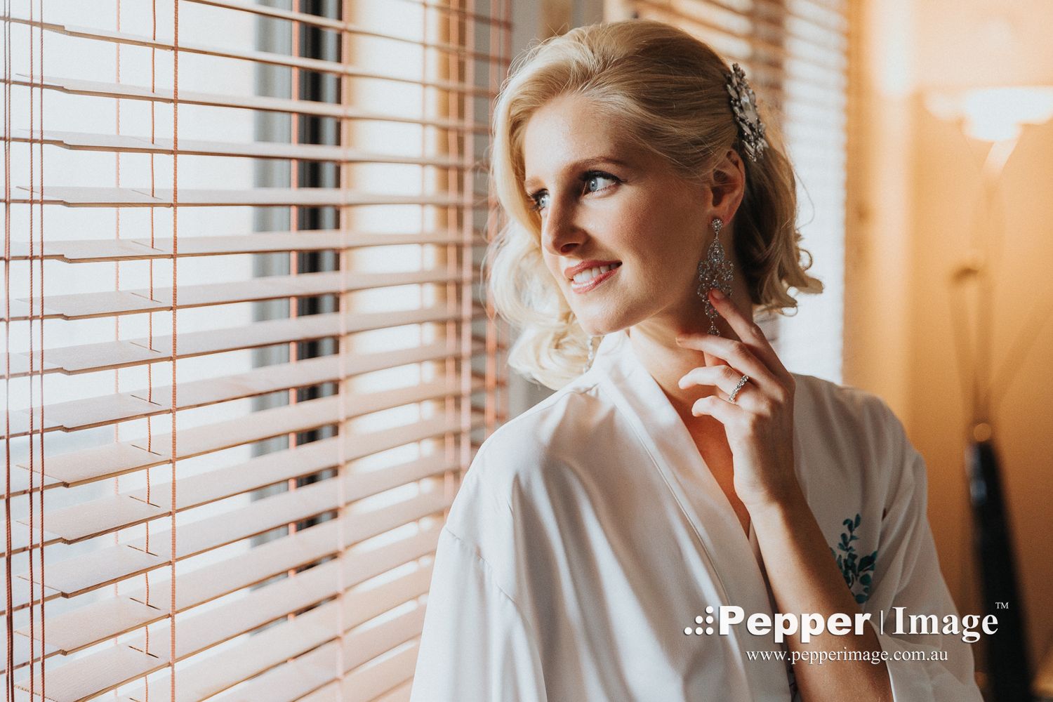 Beautiful blonde bride in white robe smiling near wooden blinds window while getting ready for wedding.