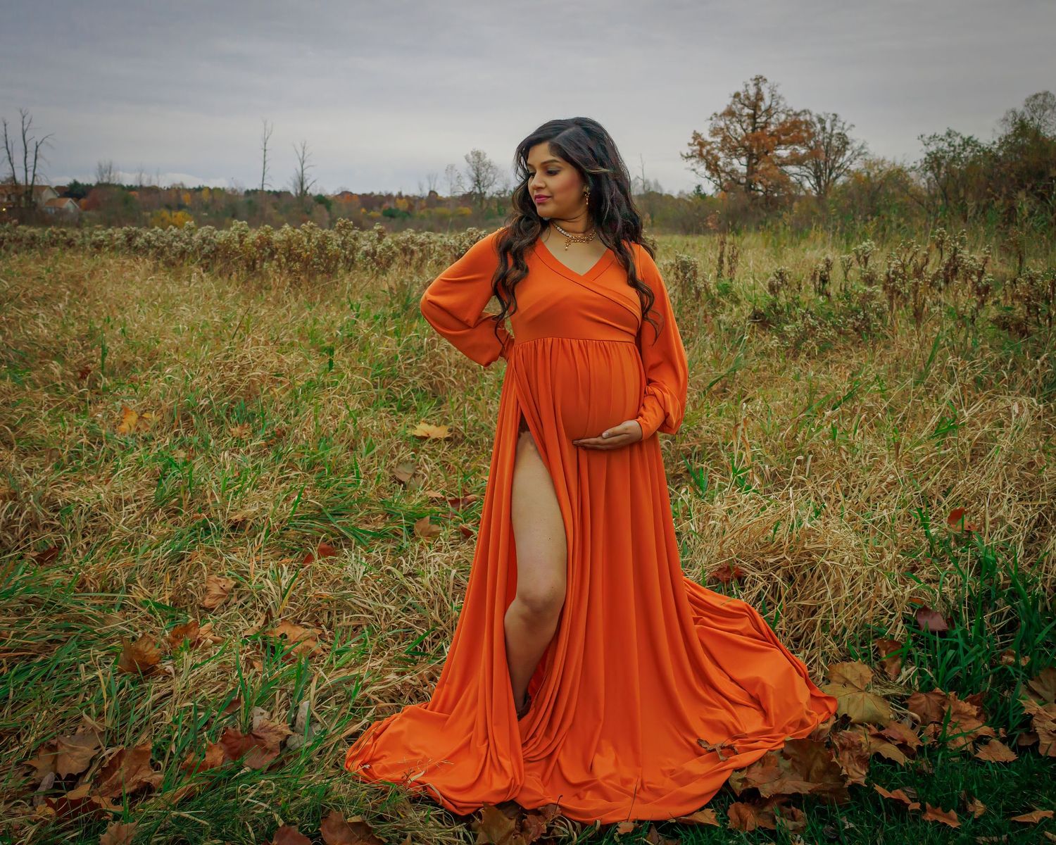 Maternity portrait in flowing orange dress against autumn field landscape.