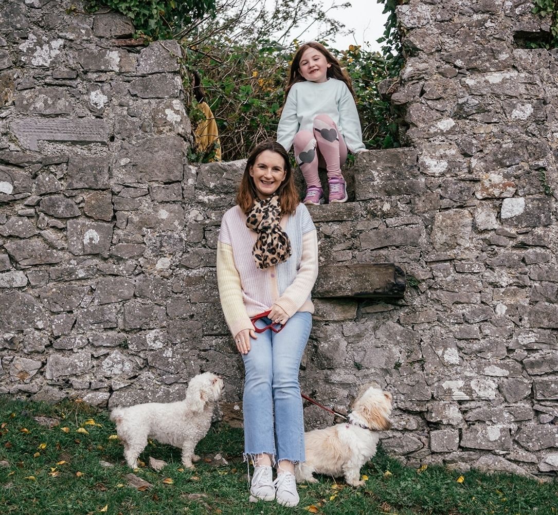 Two small white dogs pose with figures by an old stone wall.