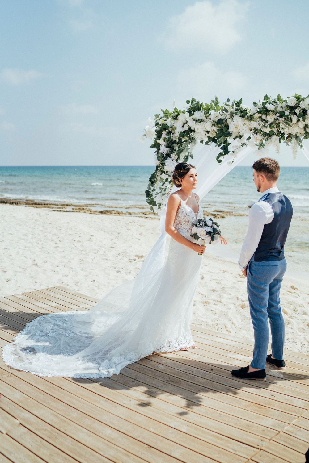 Beachfront wedding ceremony with white floral arch and wooden boardwalk.