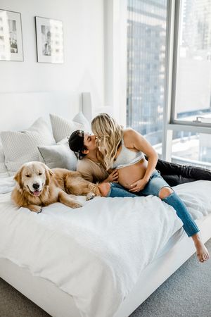 A couple relaxes on a white bed with their golden retriever in a bright modern bedroom with city views.