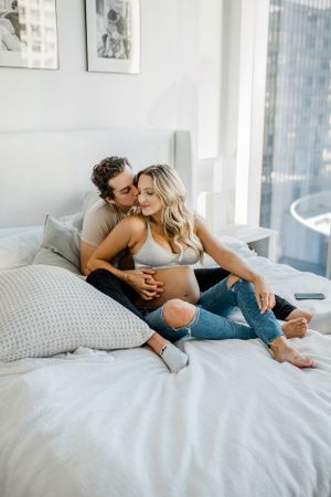 A couple lounges together on white bedding in a modern bedroom with natural light.