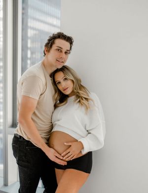 A couple poses together against a white wall with natural window light in a minimalist setting.