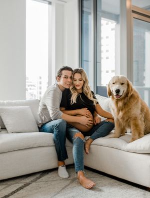 A couple relaxes on a white sectional with their smiling golden retriever in a bright modern apartment.