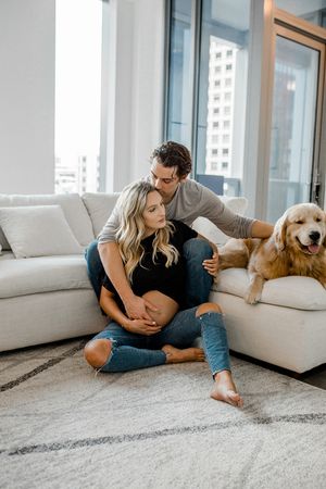 A couple and their golden retriever share a quiet moment on a white couch in a sunlit room.