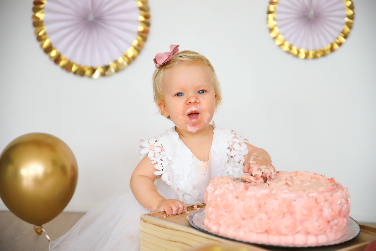 A small child in white dress sits at a table with pink cake and gold balloons for their first birthday celebration.