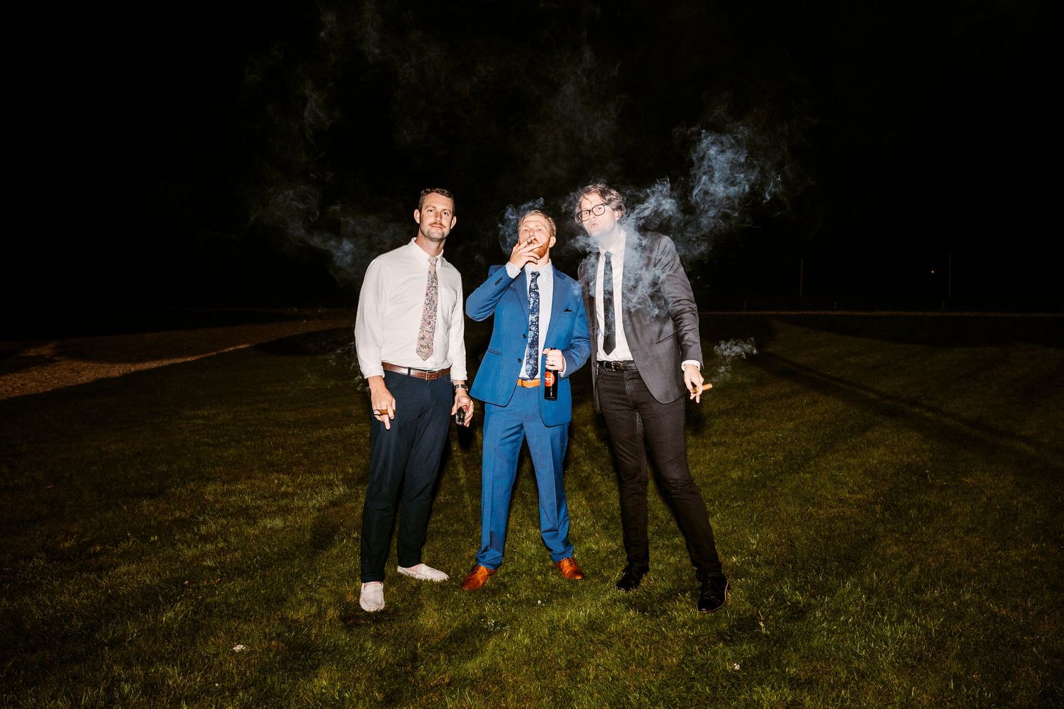 Three people sharing a lively moment at night on a grassy lawn during a wedding celebration.