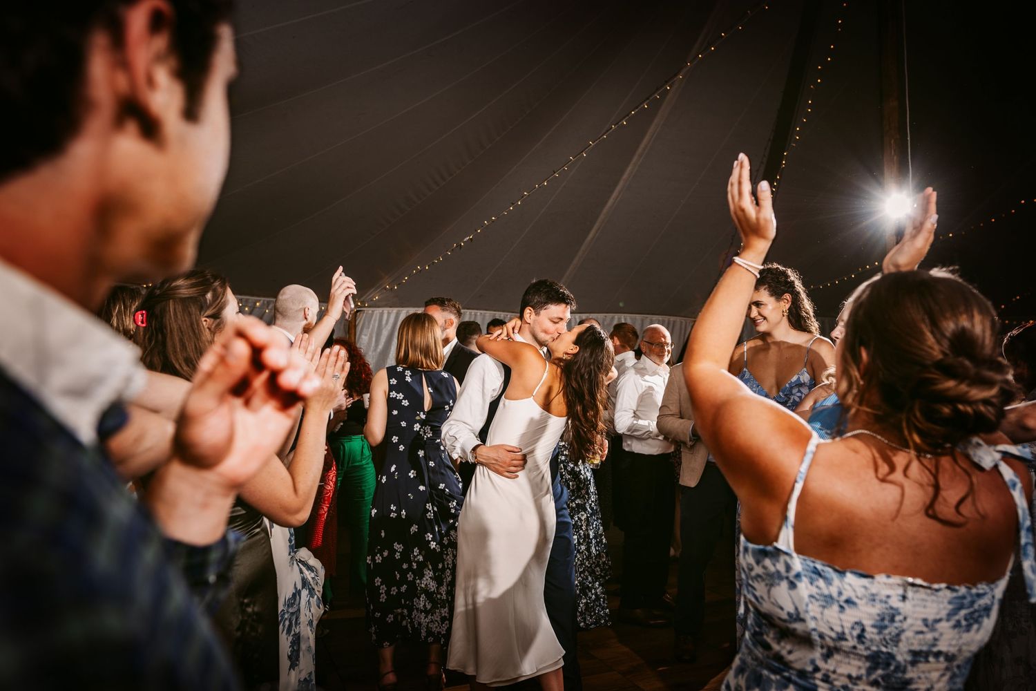 Wedding celebration with guests dancing and raising hands under illuminated tent lighting.
