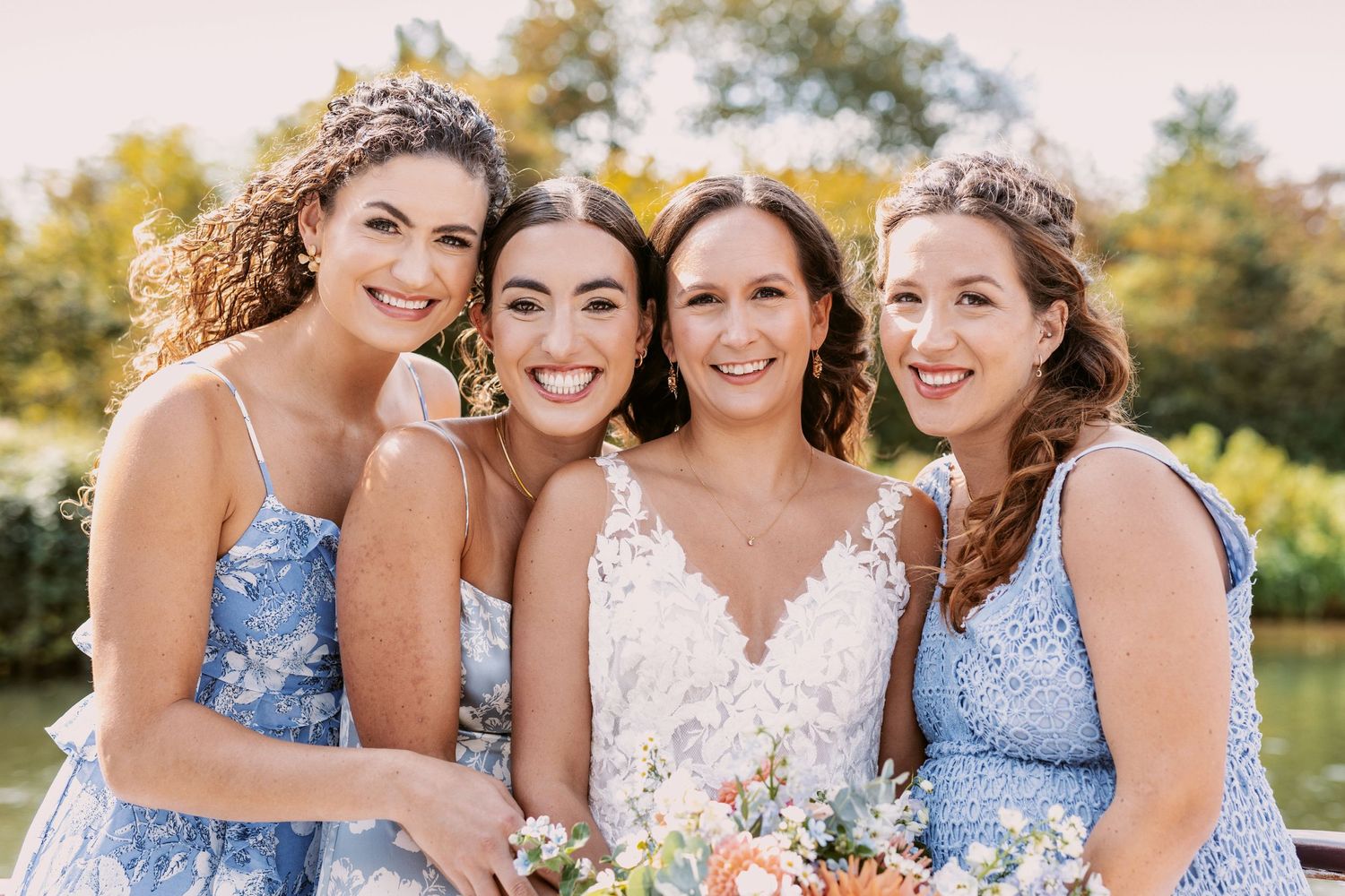 Bridal party members in light blue dresses smiling together outdoors with colorful bouquets.