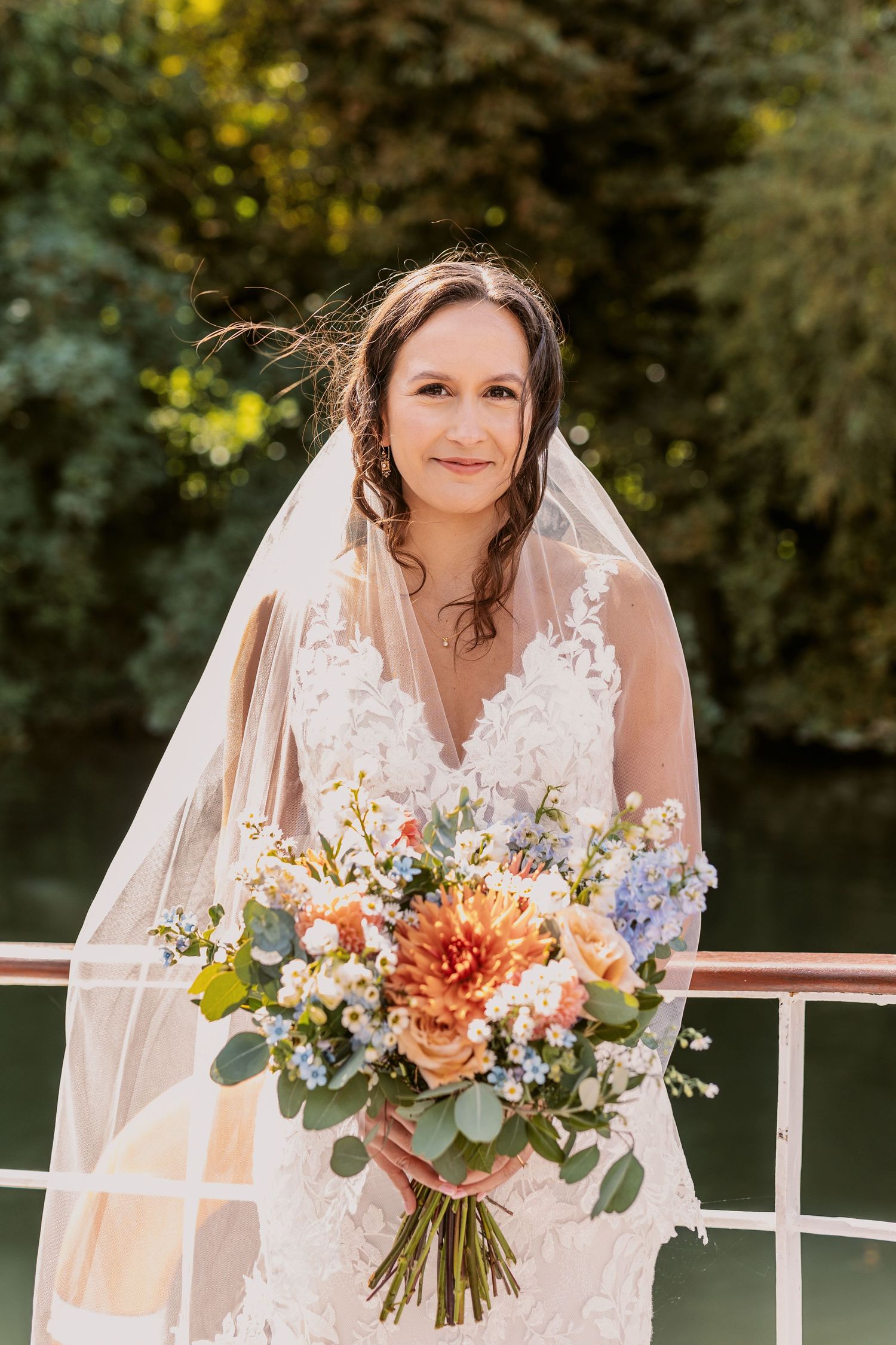 Bride in lace wedding dress holding vibrant peach and white floral bouquet outdoors.