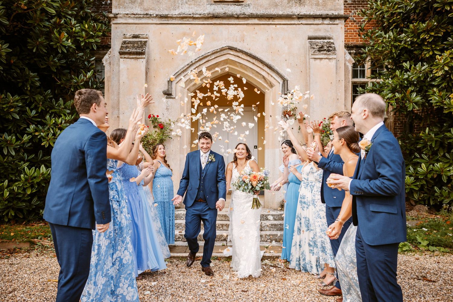 bride and groom walking through confetti as bridesmaids wearing pretty blue summer dresses and groomsmen laugh