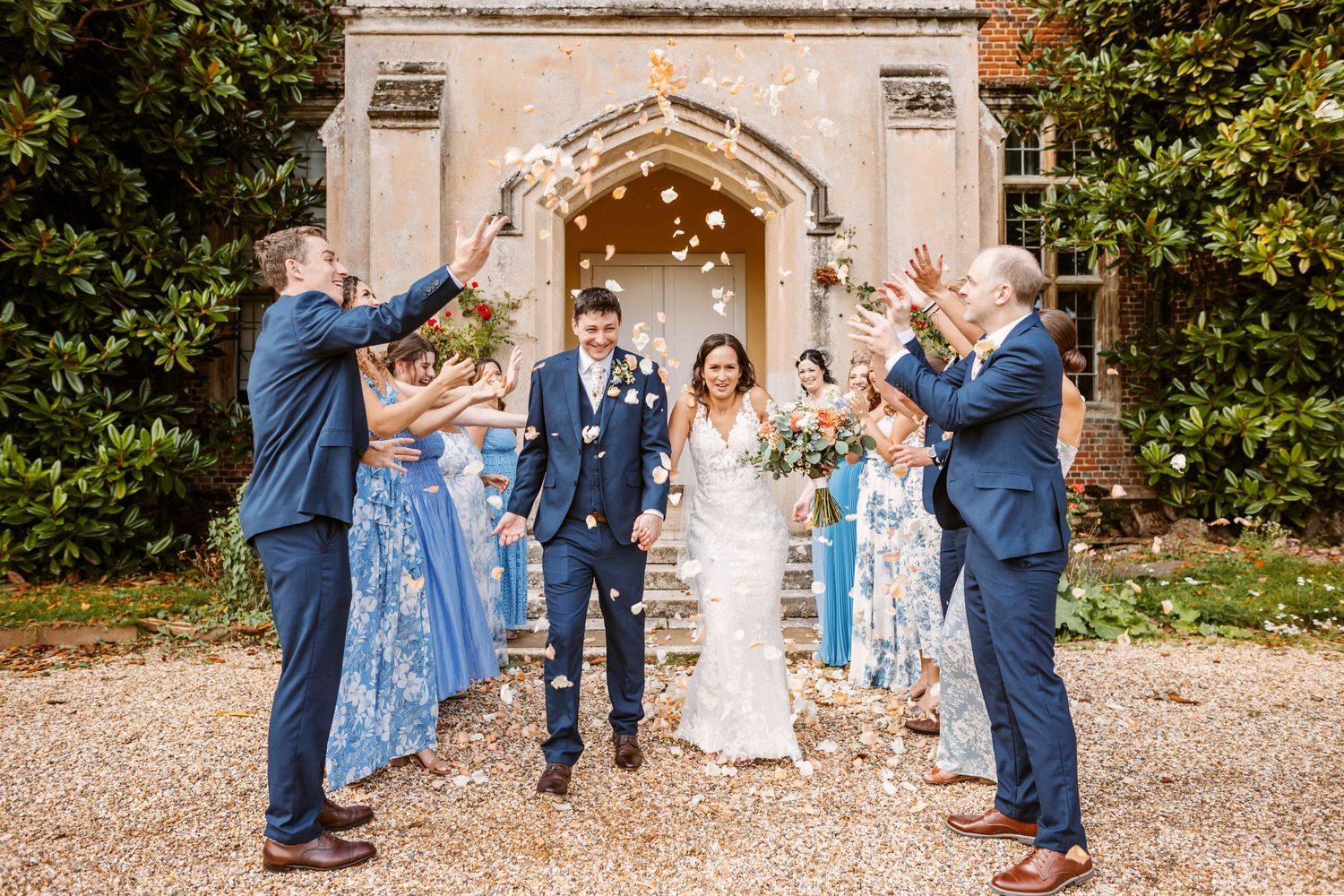 bride and groom laughing as bridesmaids wearing pretty blue summer dresses and groomsmen throw confetti over them