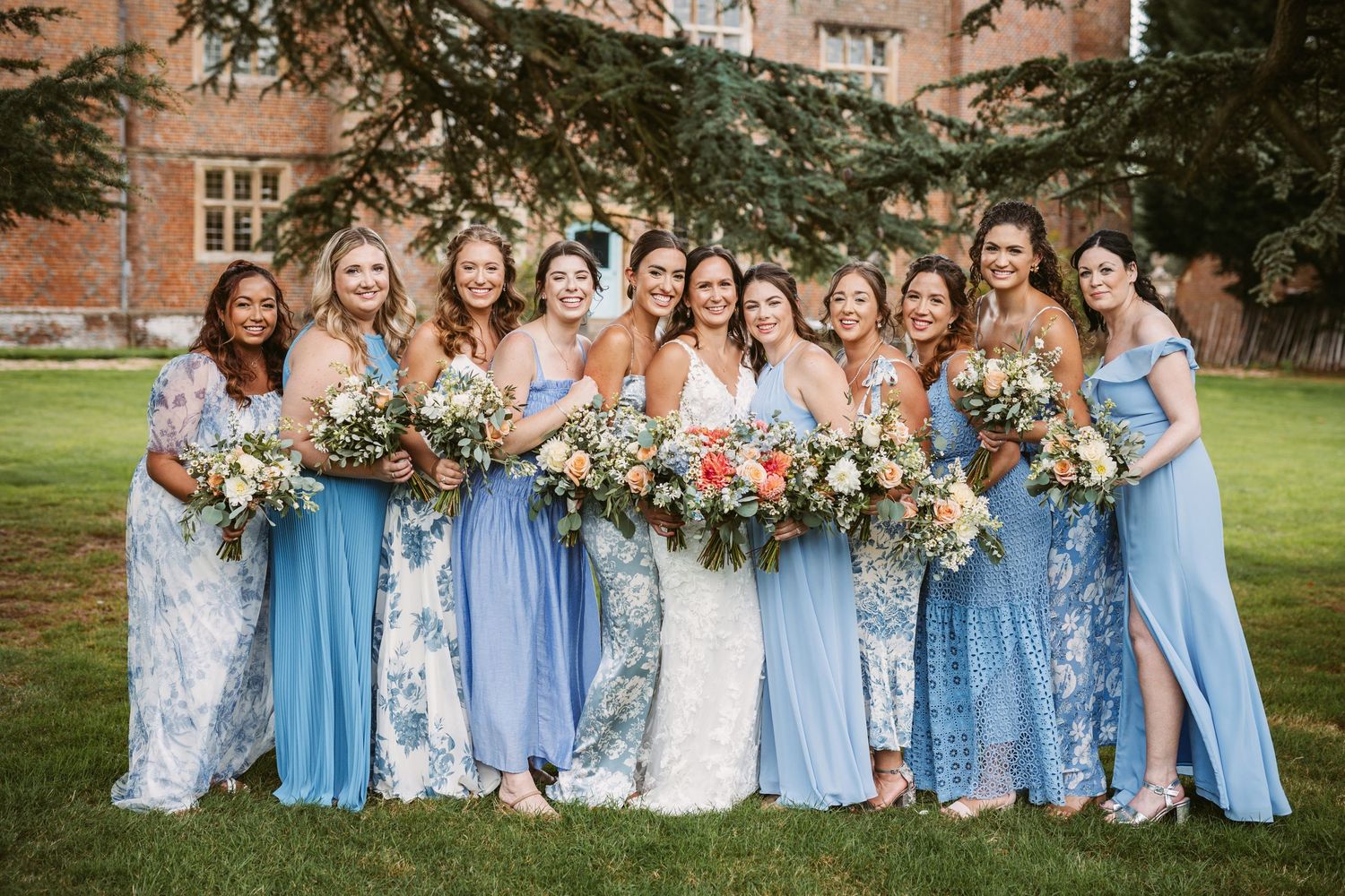 Bridal party in mix of blue and floral dresses posing with bouquets on mansion grounds.