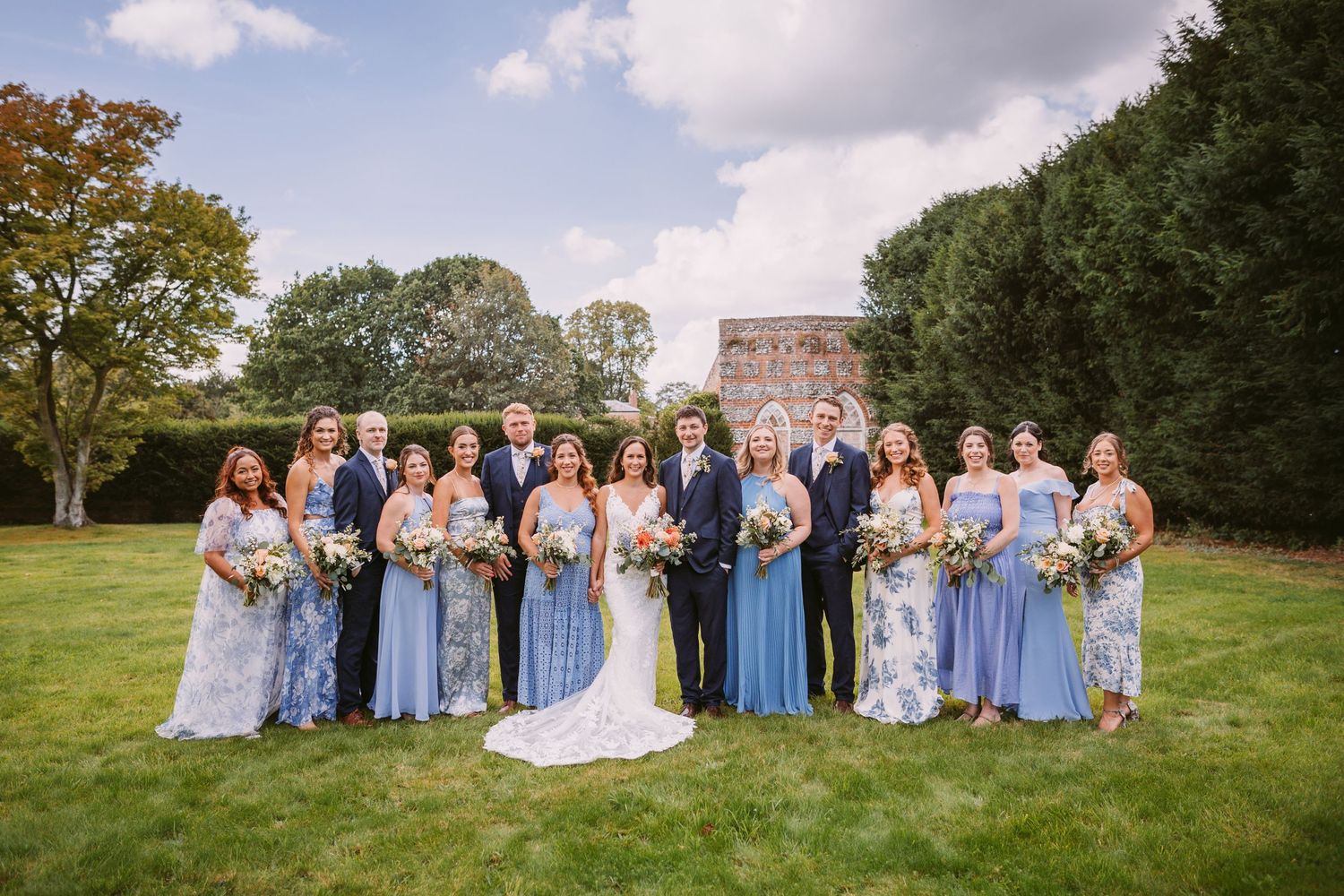 Wedding party in coordinated blue and floral dresses gathered on green lawn with brick building backdrop.