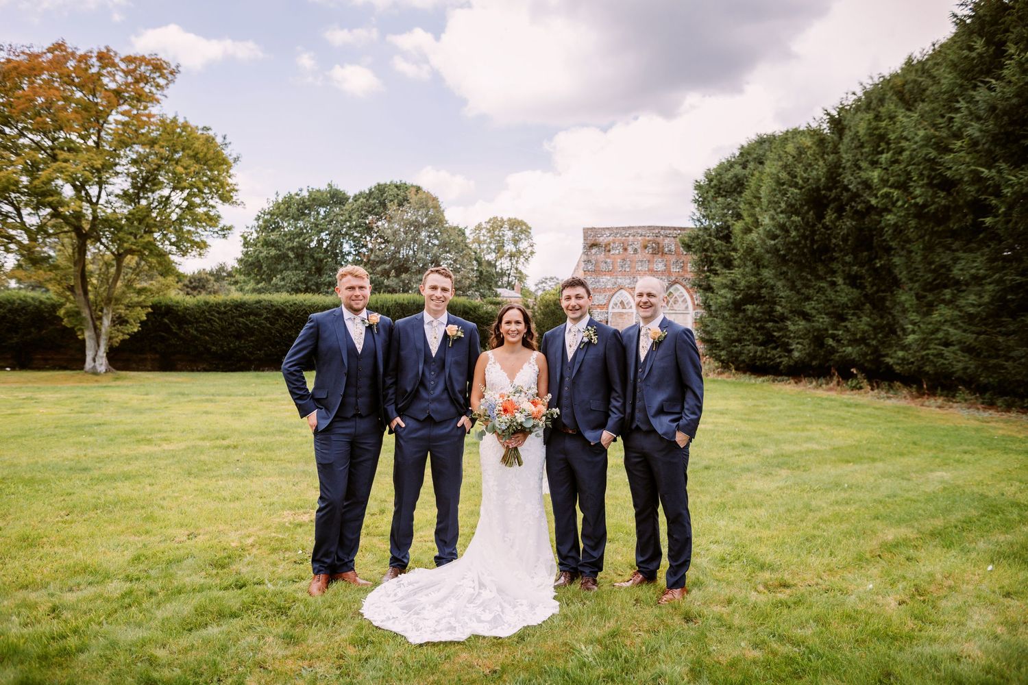 Wedding party in navy suits and white gown pose together on sunny lawn.