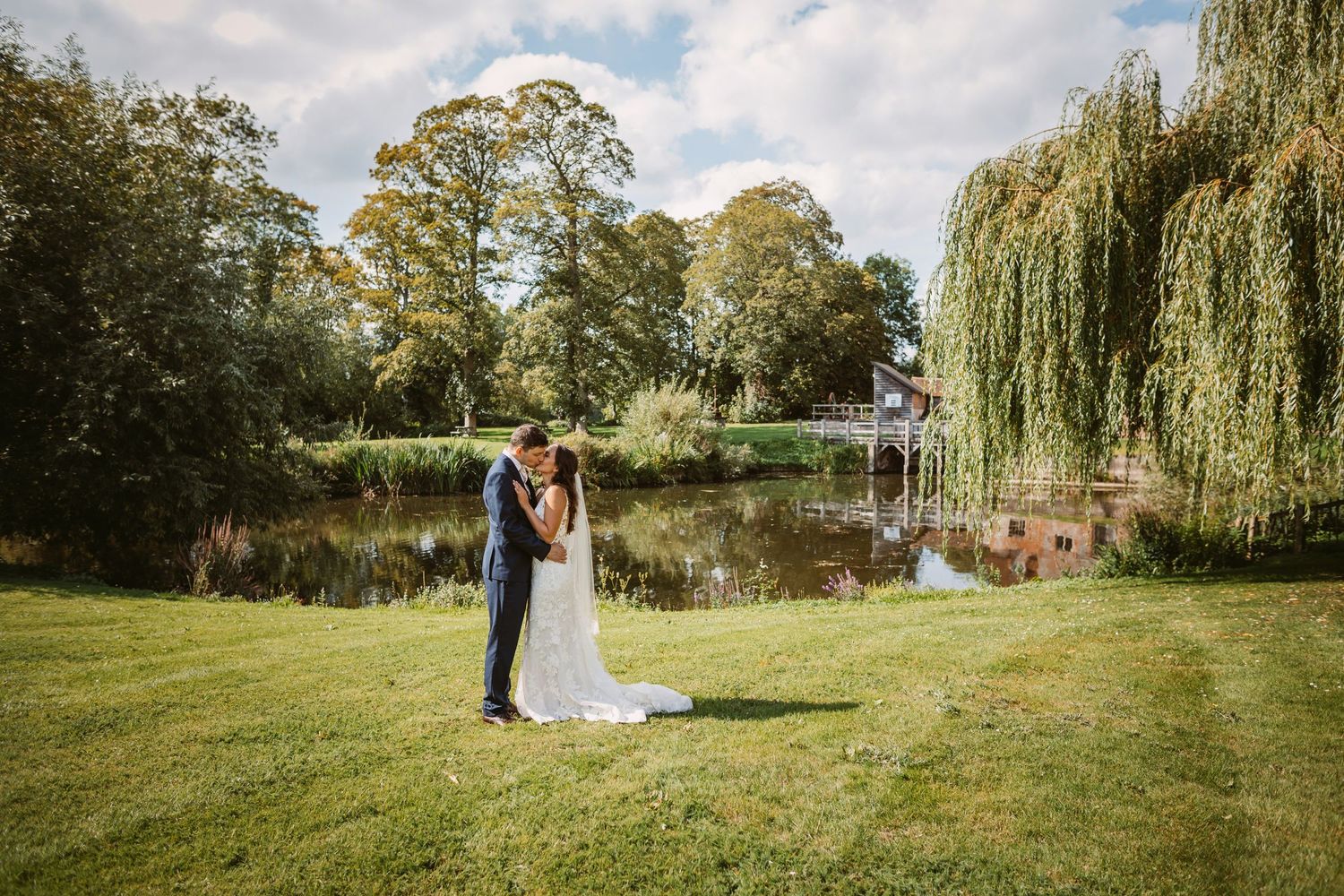 Happy young bride and groom kissing on their summery wedding day with willow trees and pond behind them