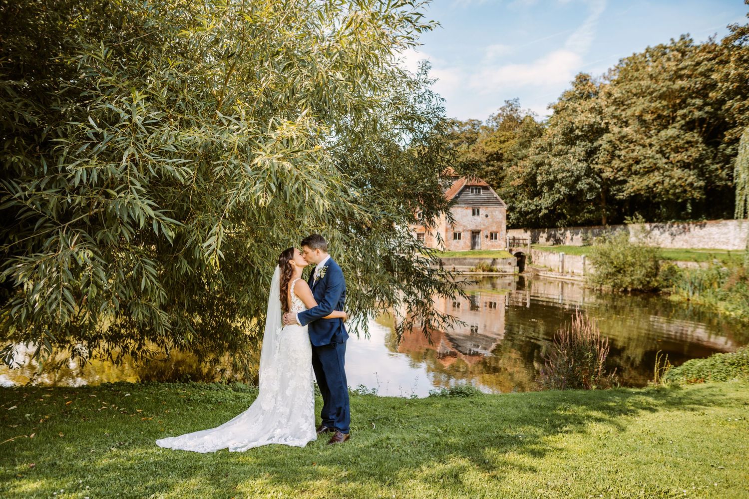 Happy young bride and groom kissing on their summery wedding day with willow trees, mill pond behind them