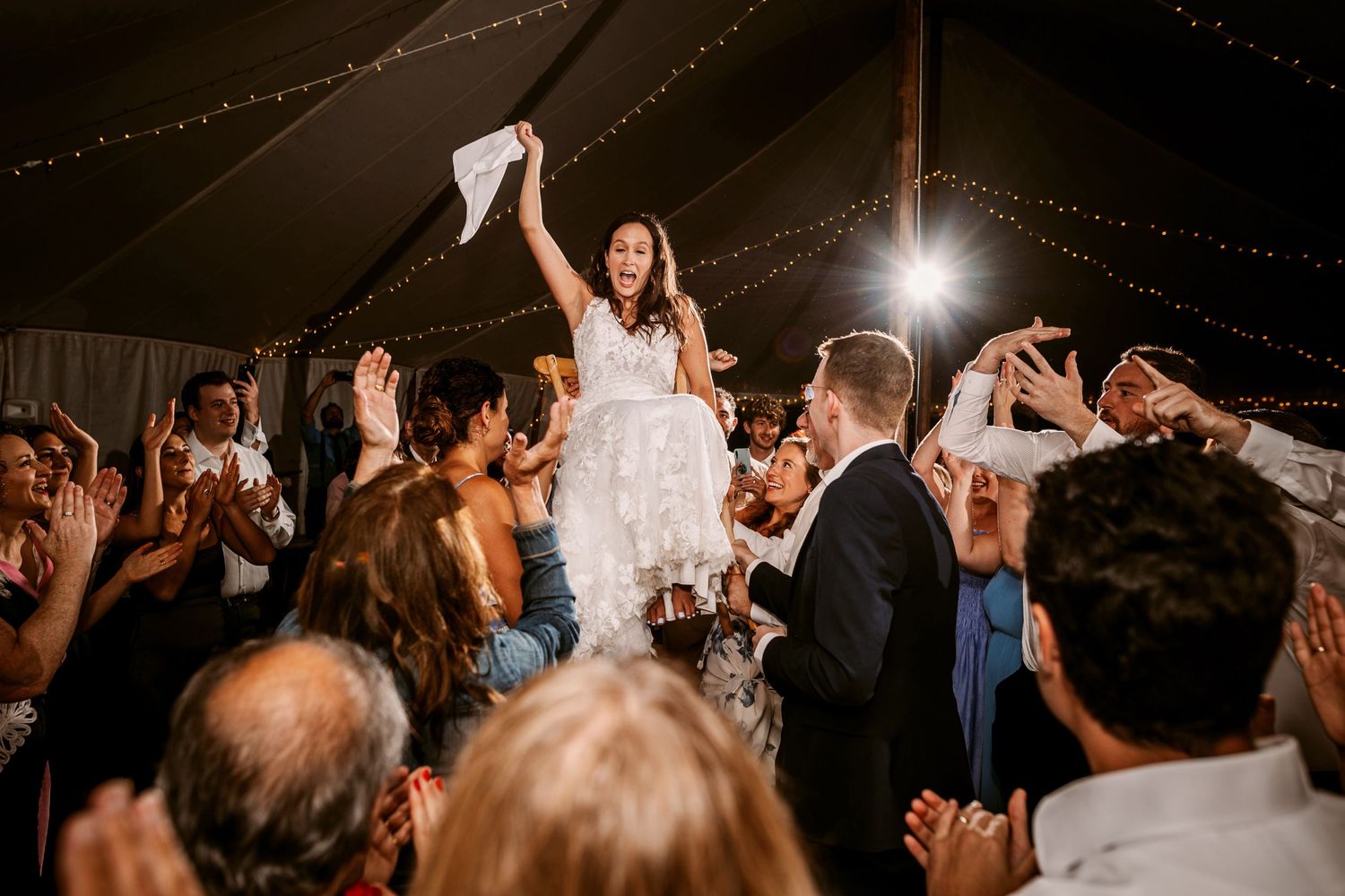 Bride lifted up on chair and dancing with friends swirling a white cloth
