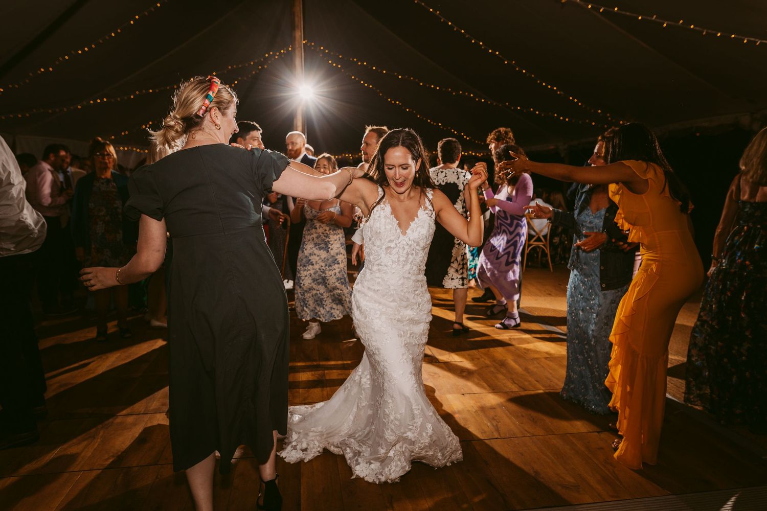 Wedding reception dance party with guests celebrating under a tent with string lights.