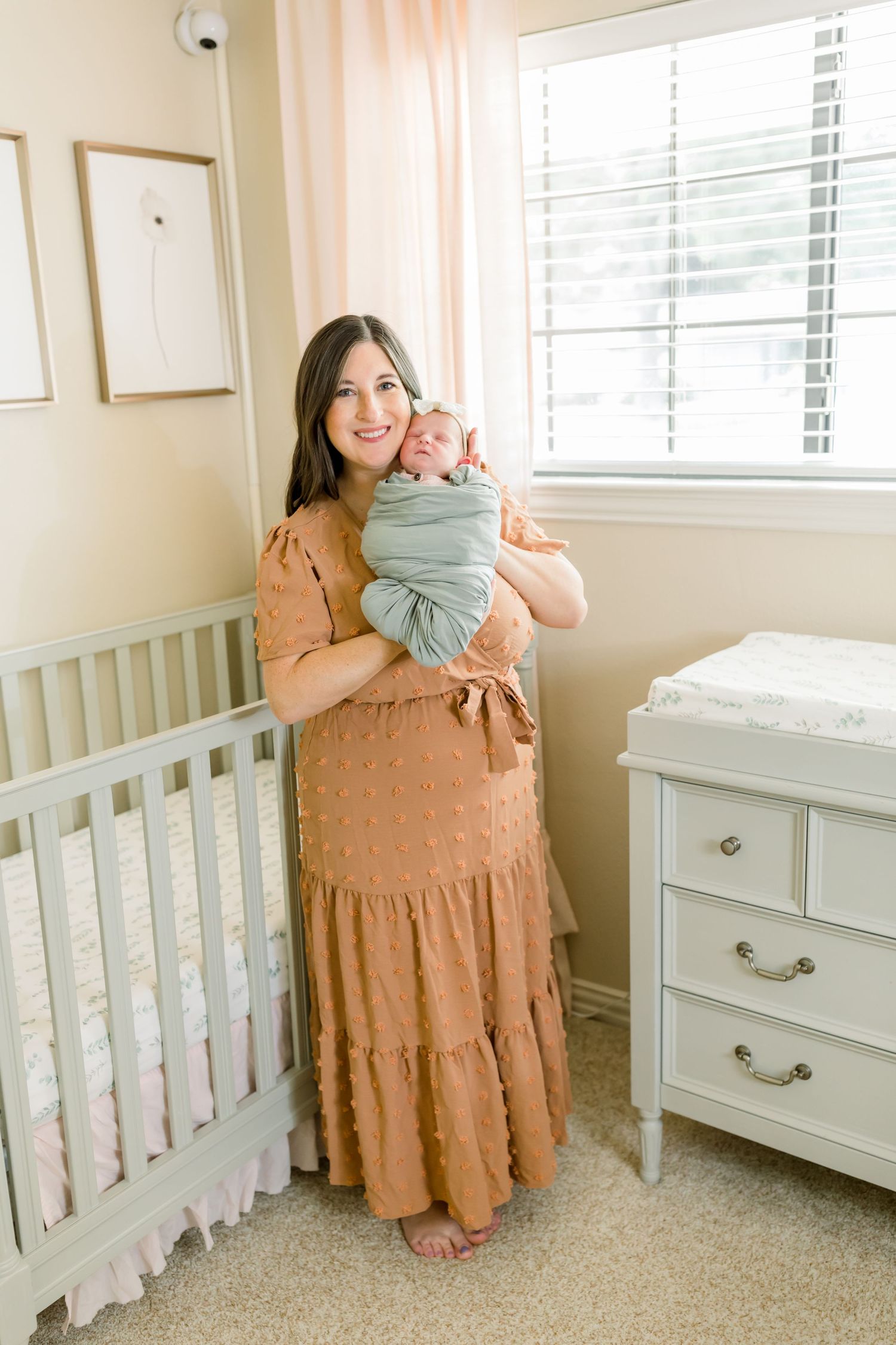 Nursery room with caregiver holding swaddled baby near white crib and dresser, wearing peach-colored dress.