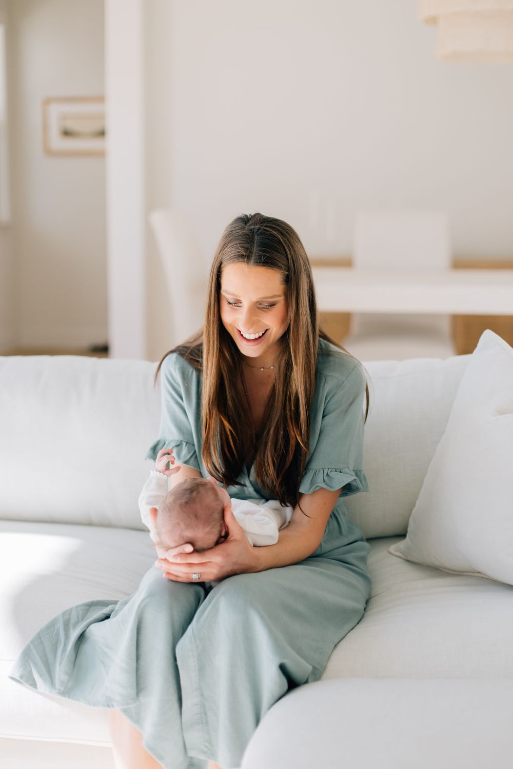 Woman in sage green dress cuddles her newborn baby while sitting on a white couch in a sunlit room.