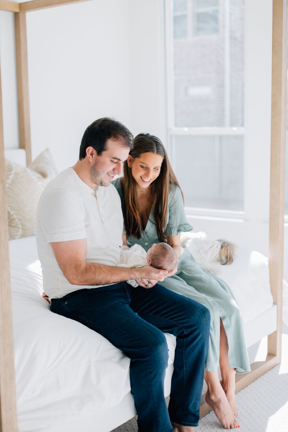 Parents sitting on a white bed share a tender moment with their newborn baby in a bright, minimalist bedroom.