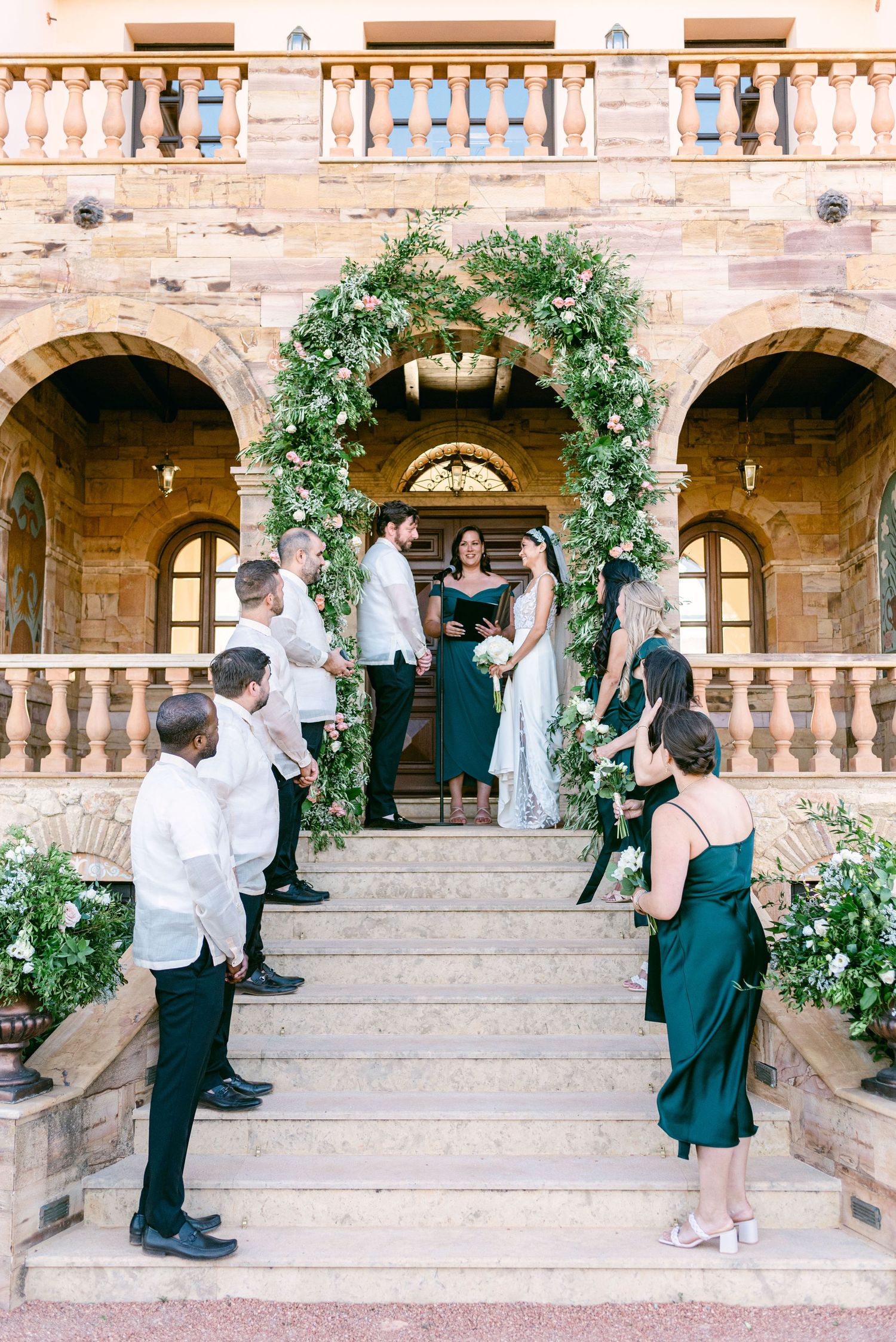A wedding ceremony under a greenery arch at a historic Mediterranean stone building.