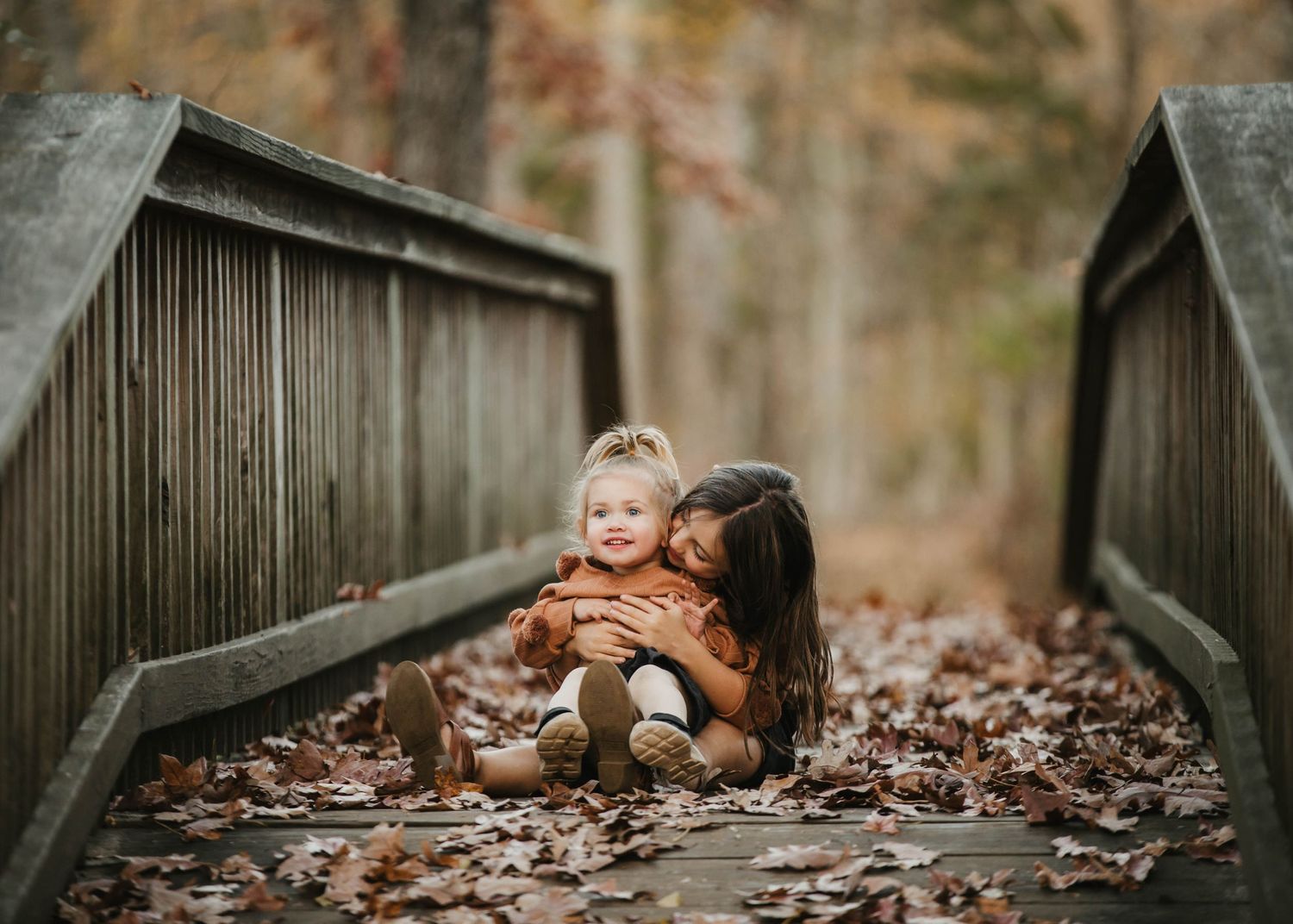Two young siblings sharing a sweet moment on a wooden bridge covered in autumn leaves in the forest.