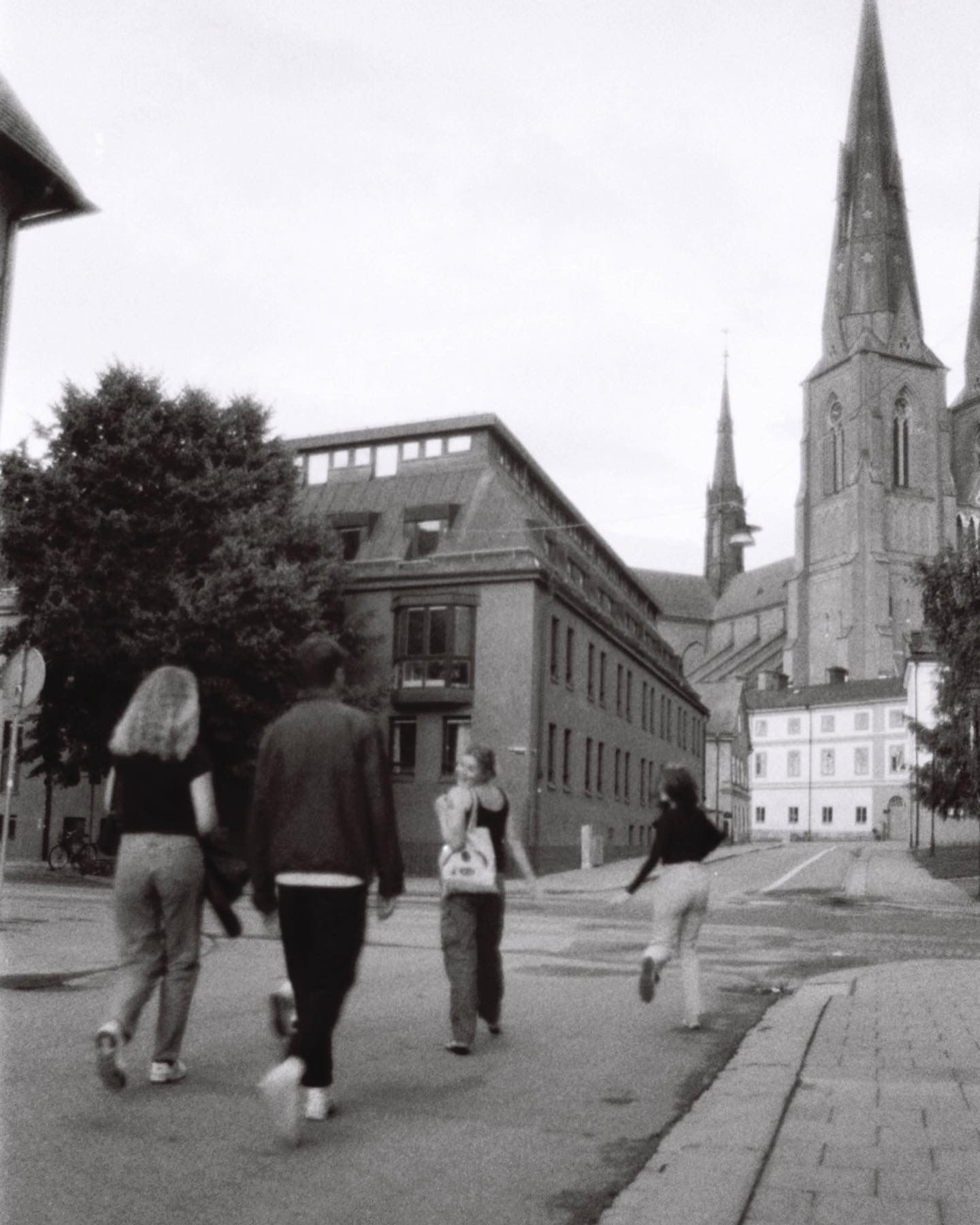 A black and white street scene showing people walking near a church spire in a European town.