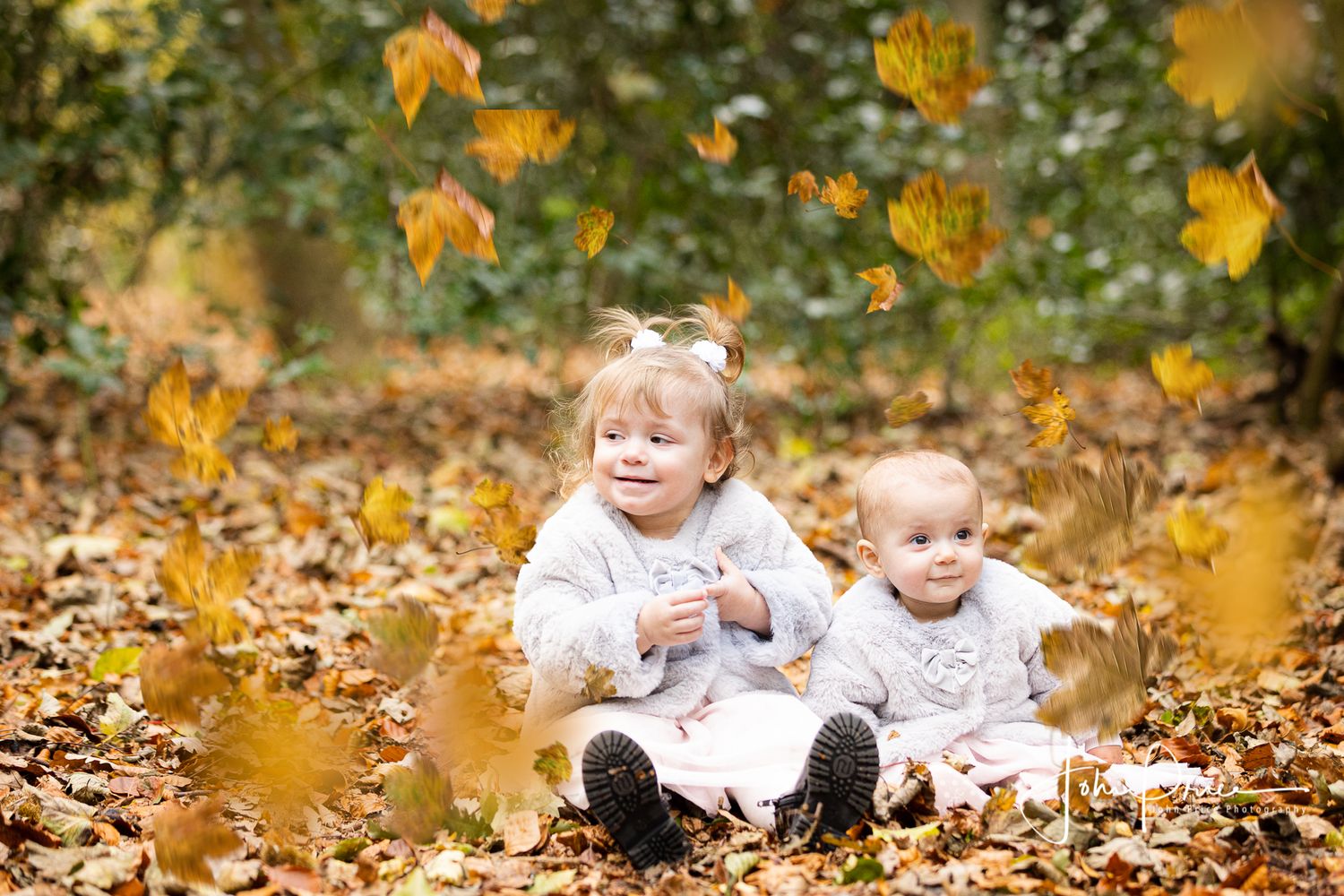 Two small children sit among fallen autumn leaves as golden foliage falls around them.