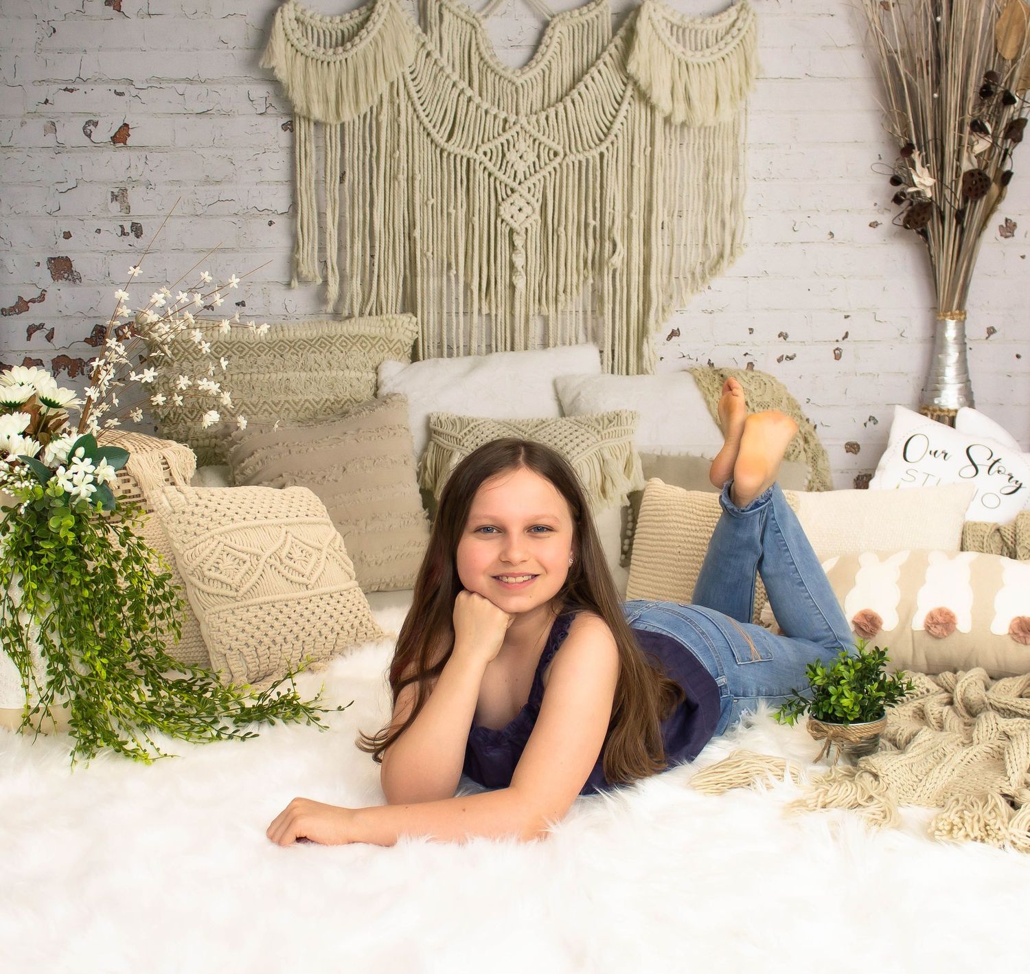 Boho-style photo setup with macrame wall hanging, throw pillows, plants and white textured backdrop.