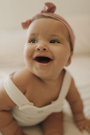 Joyful baby wearing a pink headband and white outfit smiles brightly at the camera.