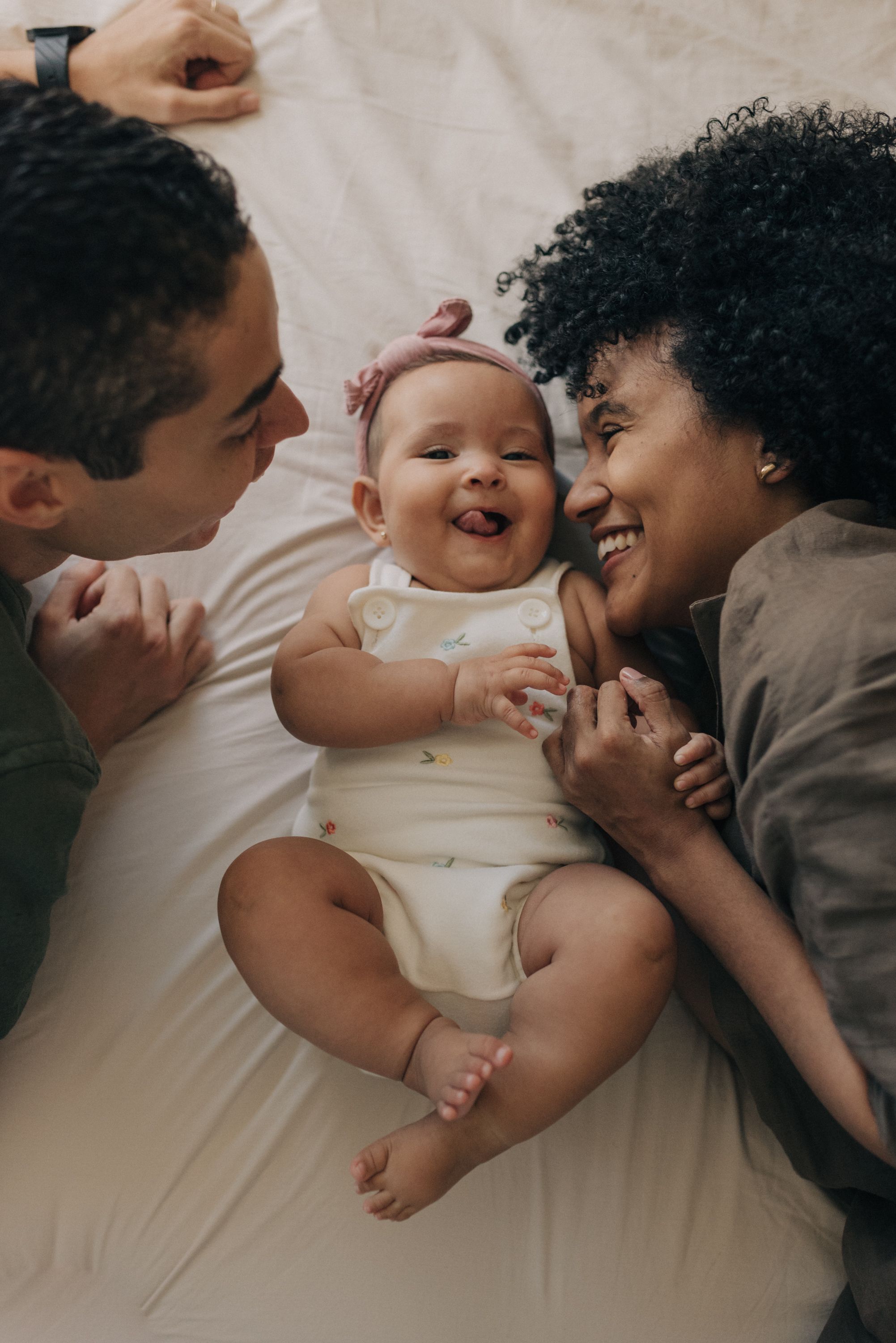 Happy family moment with baby laughing while laying between parents on white bedding.