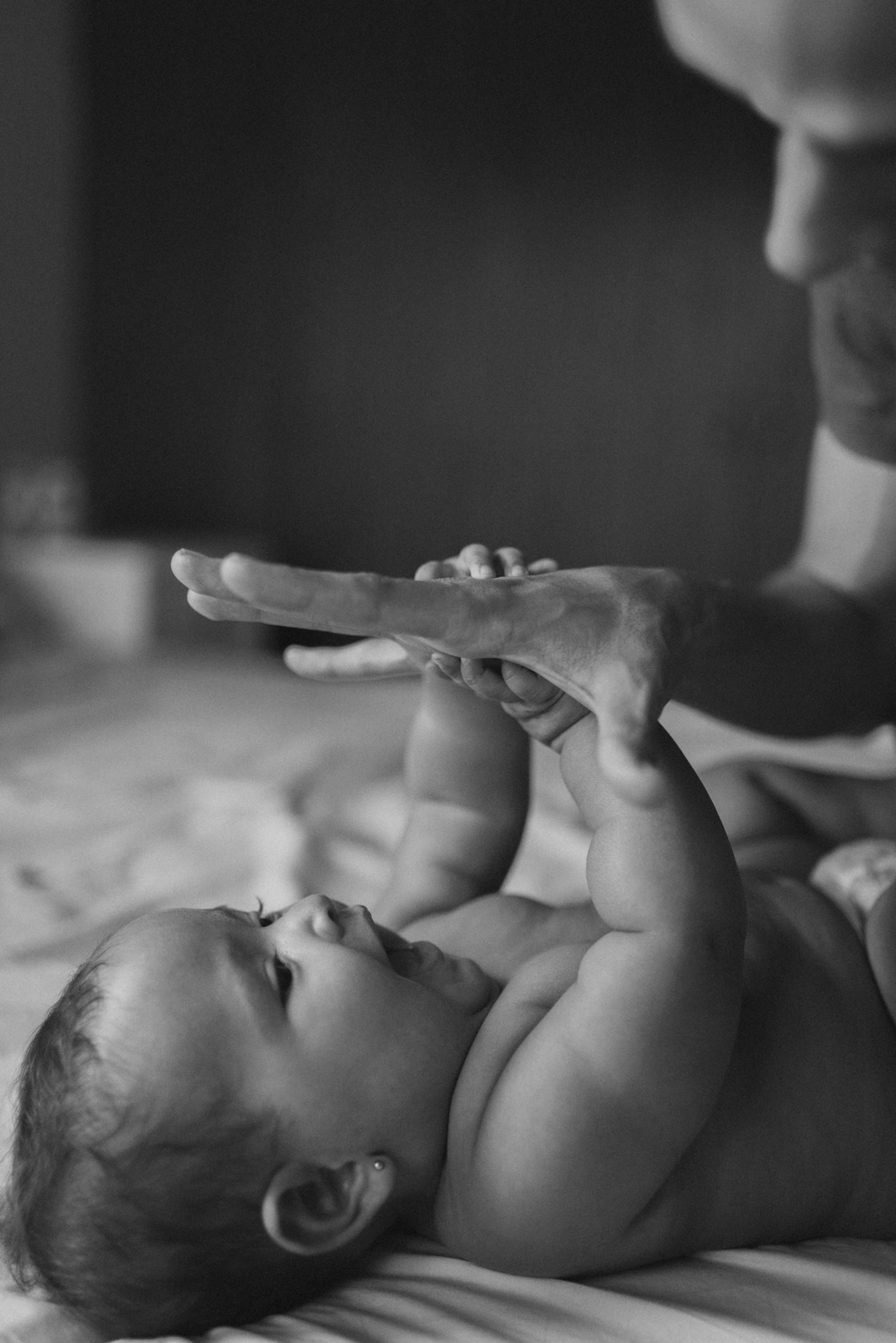 Black and white image of baby reaching up to touch adult's hand while lying on bed.