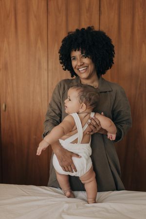 Baby in white romper stands supported while being held from behind on bed.