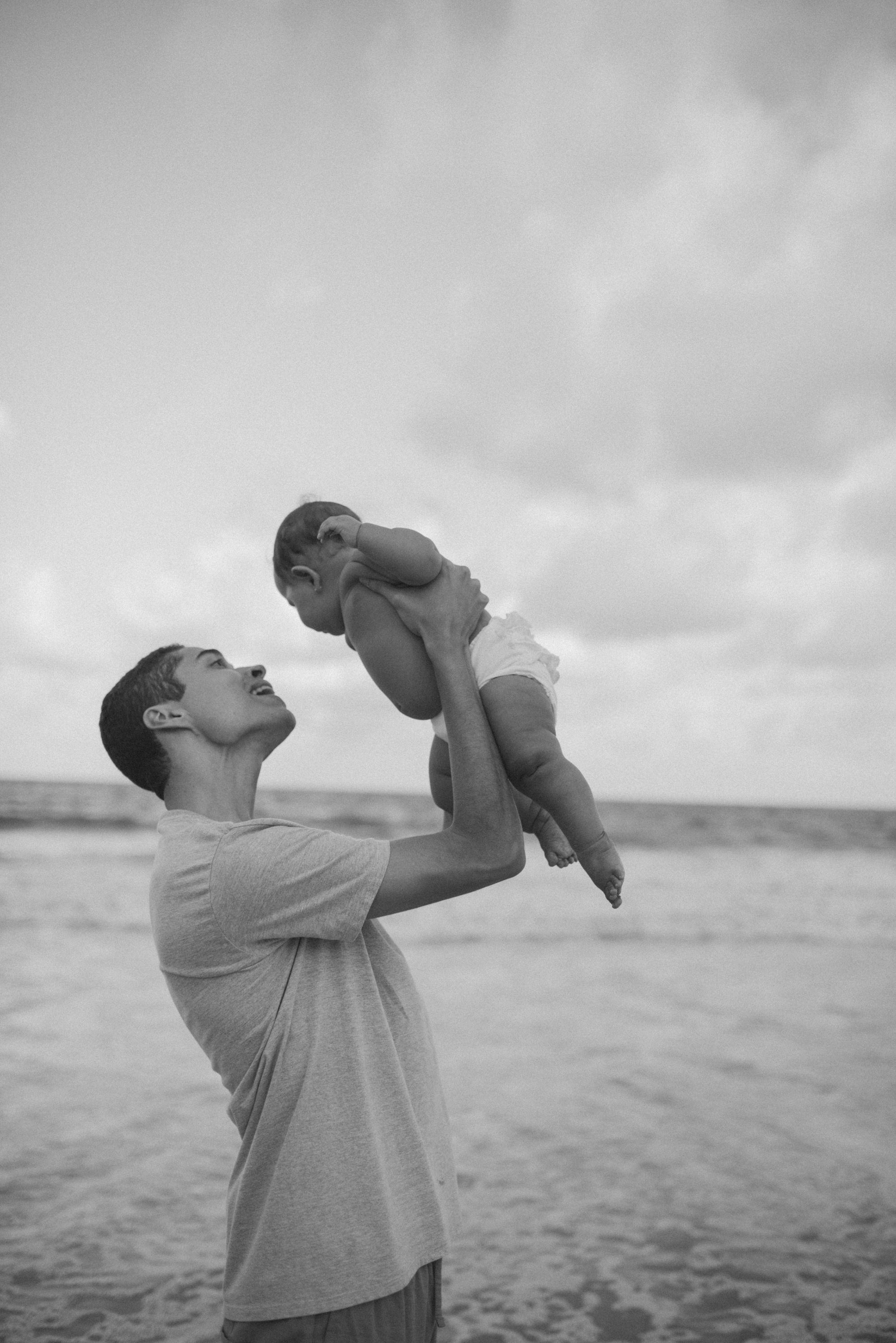 Black and white beach scene of adult lifting baby against cloudy sky.