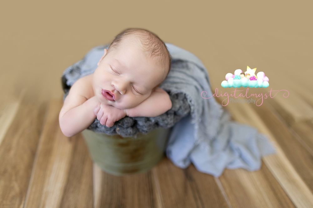 Sleeping baby posed in gray bucket on wooden surface.