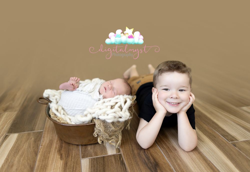 Two siblings on wooden floor with one resting in rustic bucket.