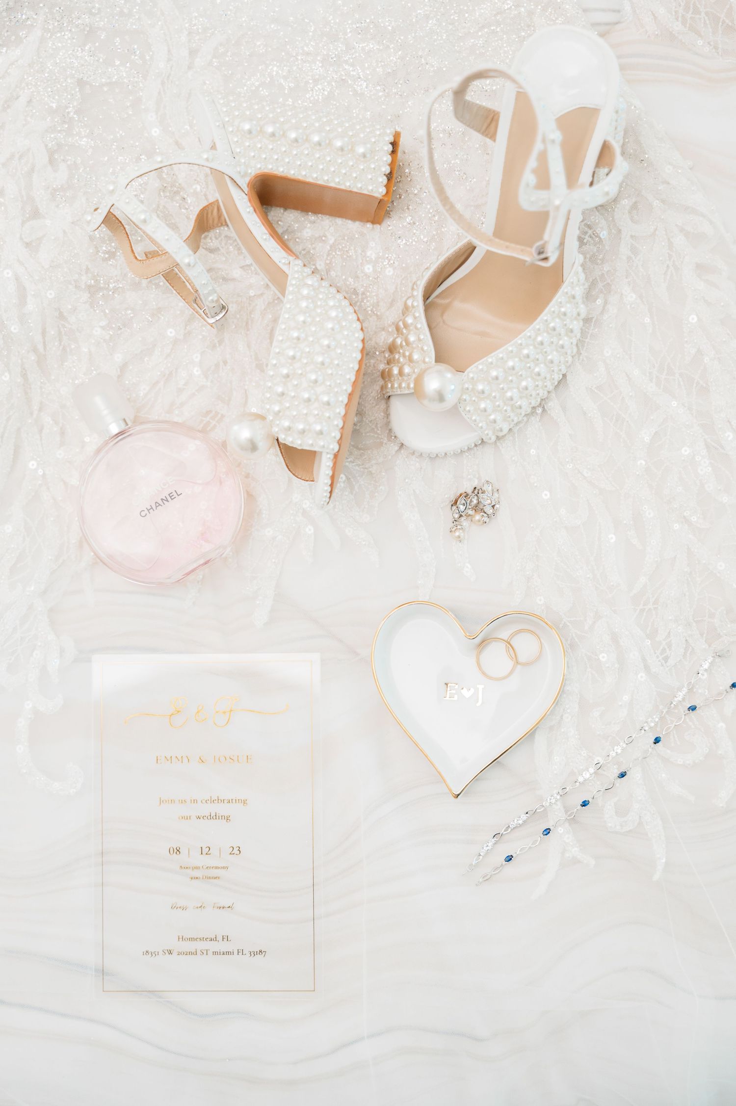 Wedding details flatlay with pearl shoes, invitation, and heart decoration on white fabric.