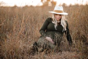 An expectant woman in a dark sweater and white hat sits in a golden field at sunset.