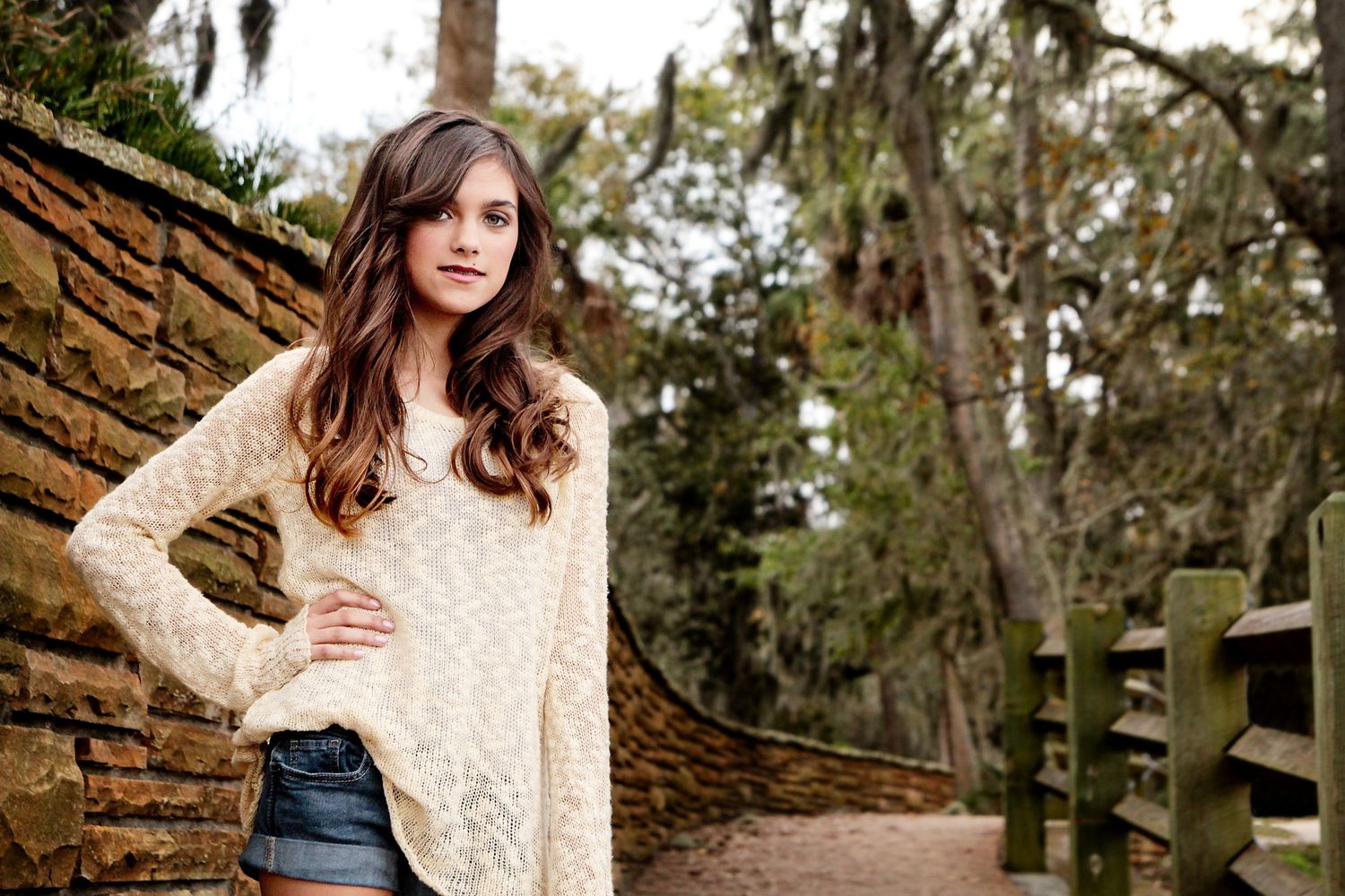 Teenage girl in cream colored shirt with jean shorts posing by a brick wall at Phillipe Park in Safety Harbor.