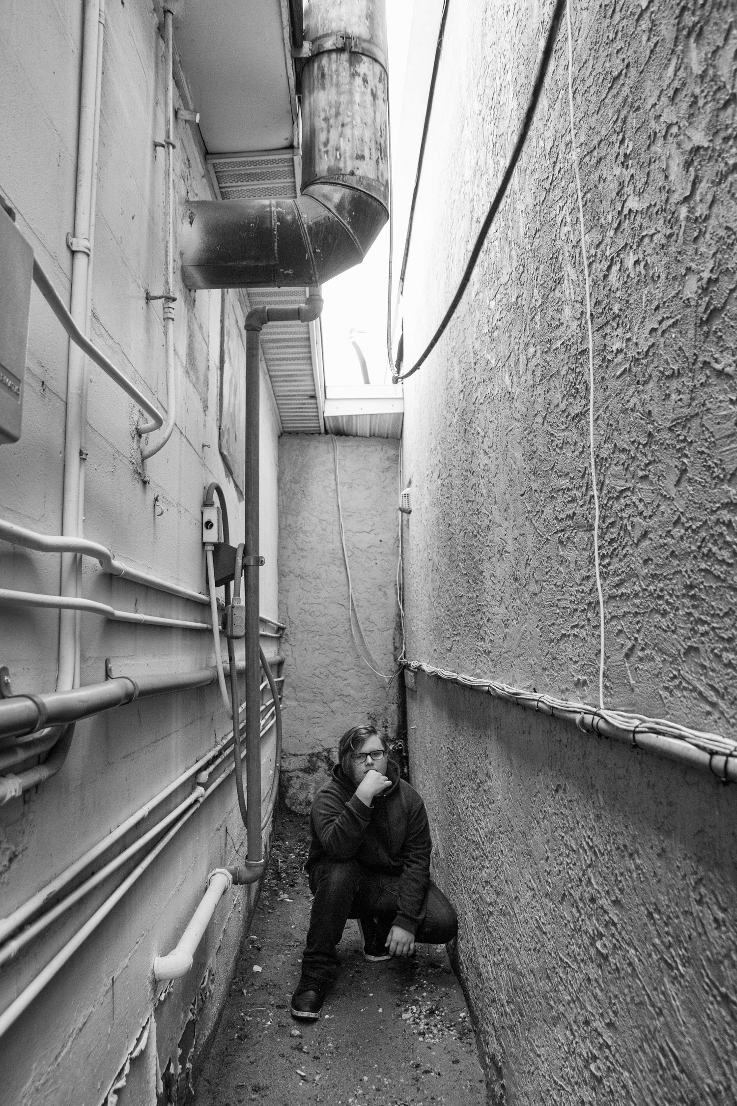 Teenage boy wearing all black, crouched in an alleyway.