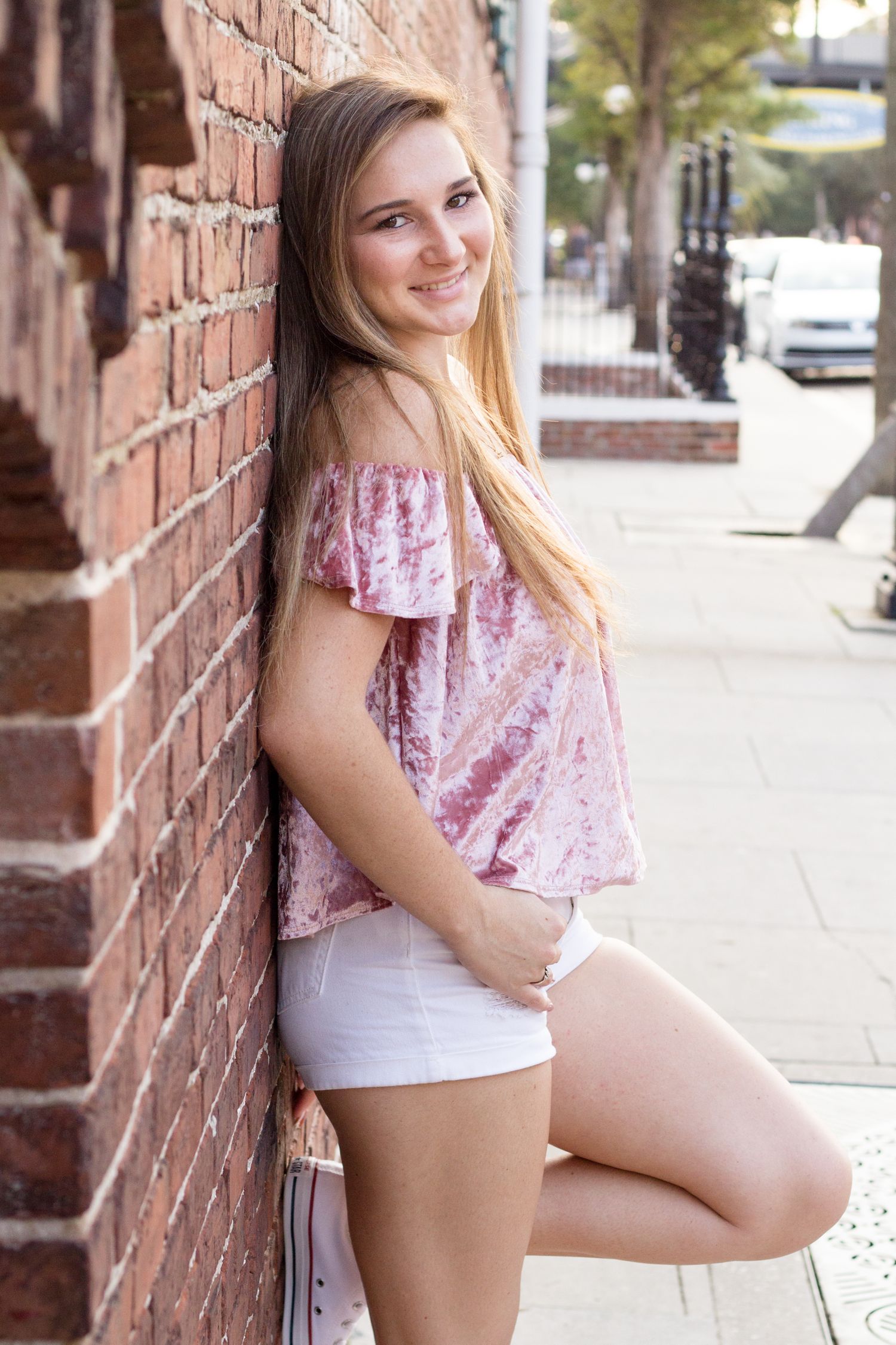 Teenage girl in pink shirt and white shorts posing against brick wall.
