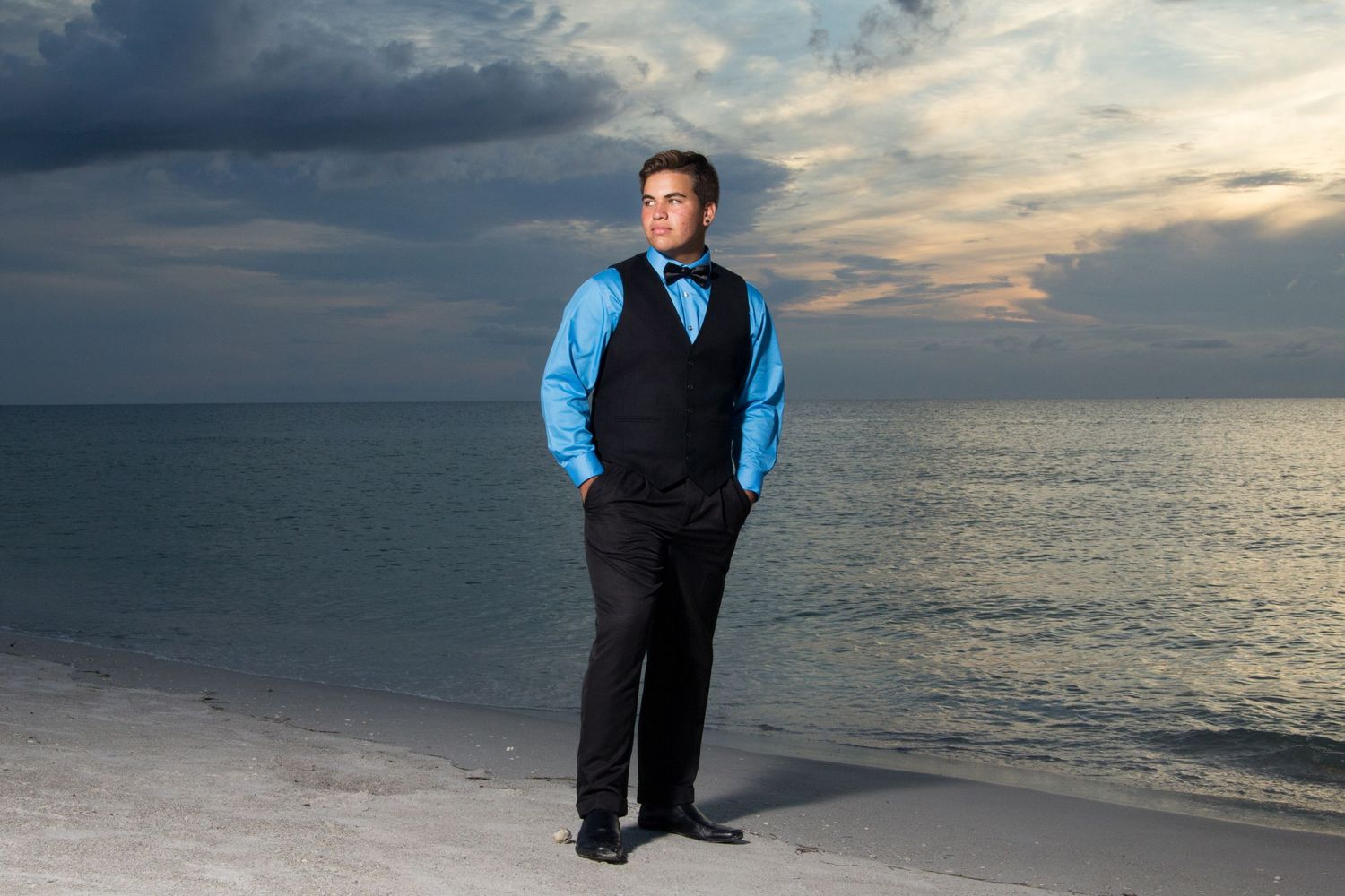 Young man in a blue dress shirt with black vest and pants posing at the sea shore at sunset.